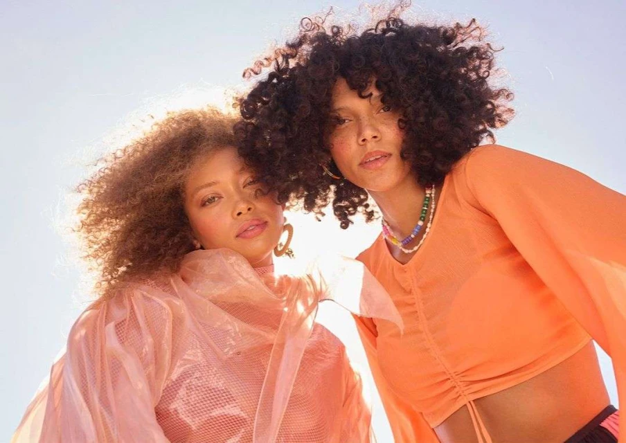 Two women with curly hair pose outdoors in peach and orange tops against a bright sky, showcasing natural beauty and fashion.