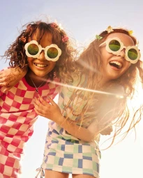 Two young girls wearing oversized flower-shaped sunglasses and checkered dresses smiling outdoors in bright sunlight.