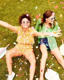 Two smiling women in sunglasses lying on grass surrounded by falling colorful confetti with wooden paddles nearby