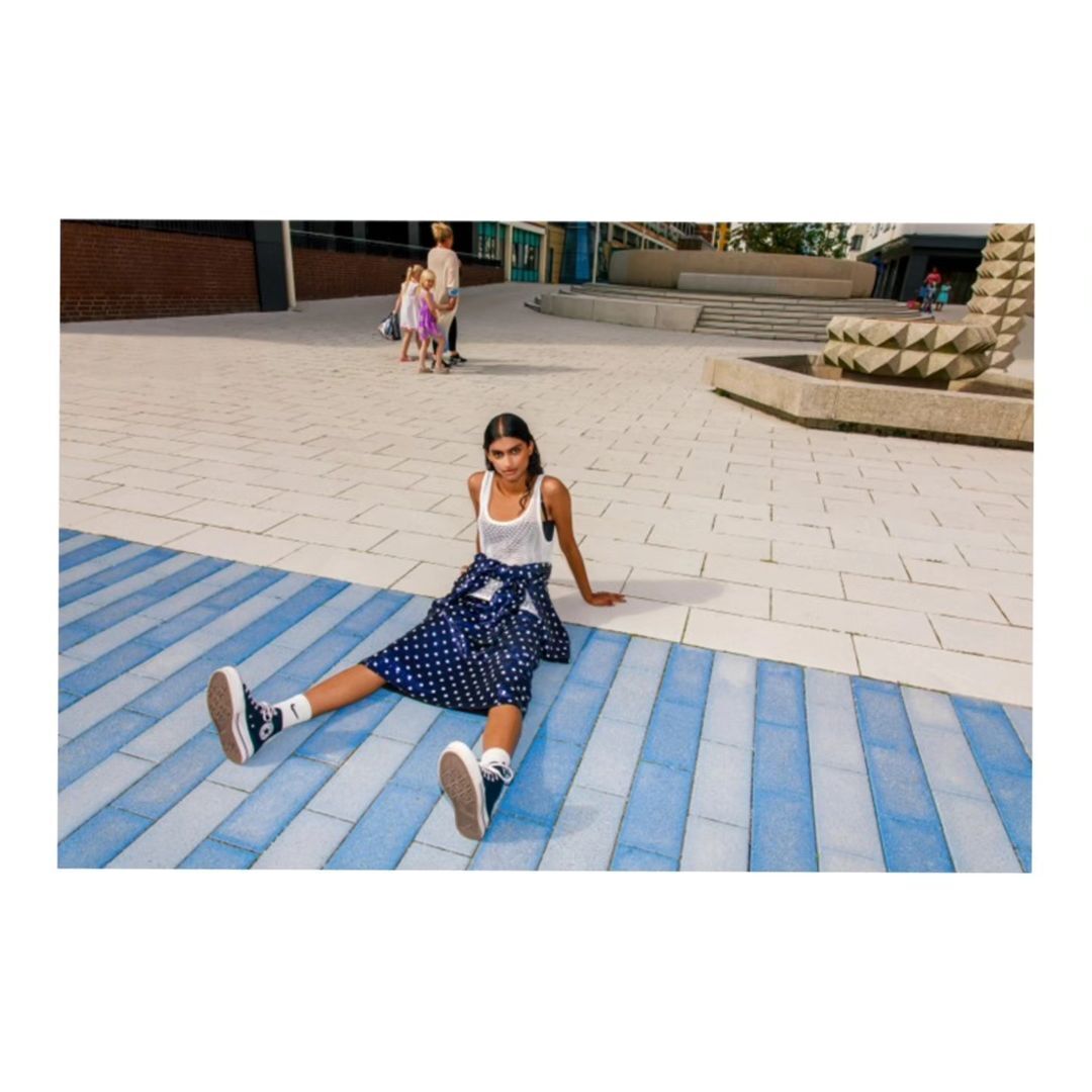 Woman in tank top and polka dot skirt sitting on striped pavement. People and modern architecture in background. Fashion shoot outdoors.