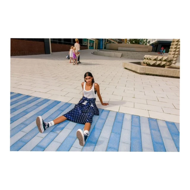 Woman in tank top and polka dot skirt sitting on striped pavement. People and modern architecture in background. Fashion shoot outdoors.