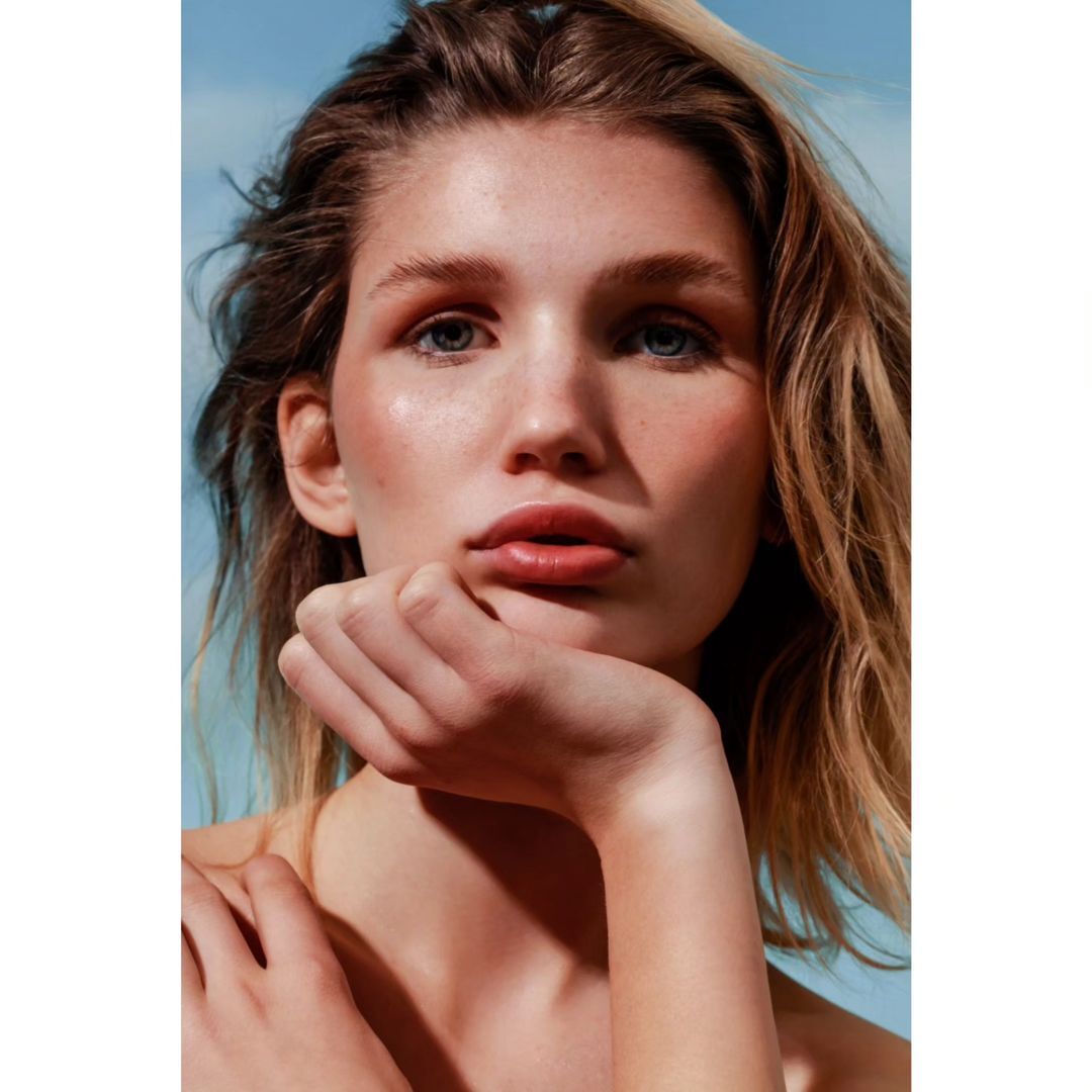 Close-up portrait of a woman with fair skin and light brown hair, looking directly at the camera against a soft blue background.