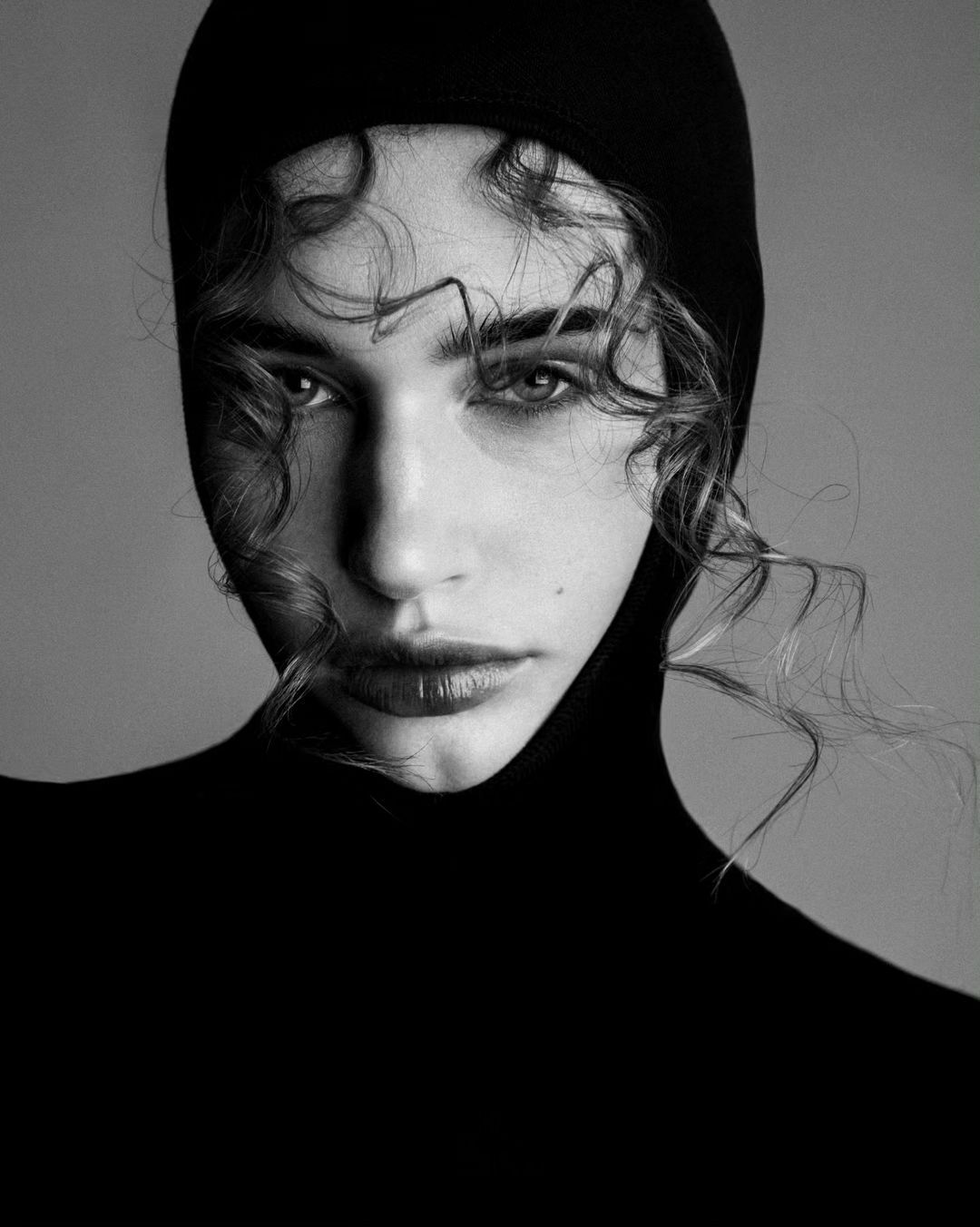 Striking monochrome portrait of a young woman with curly hair, wearing a black hood, showcasing dramatic lighting and sleek beauty.