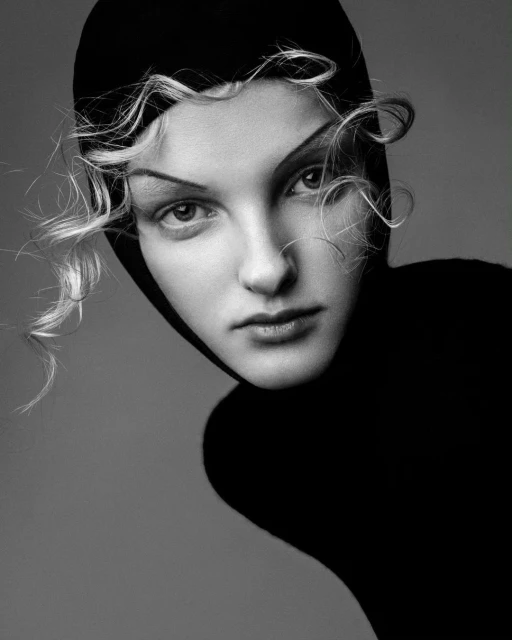 Monochrome close-up portrait of a woman with fair skin wearing a dark hood, showcasing a minimalist aesthetic and curly blonde hair.