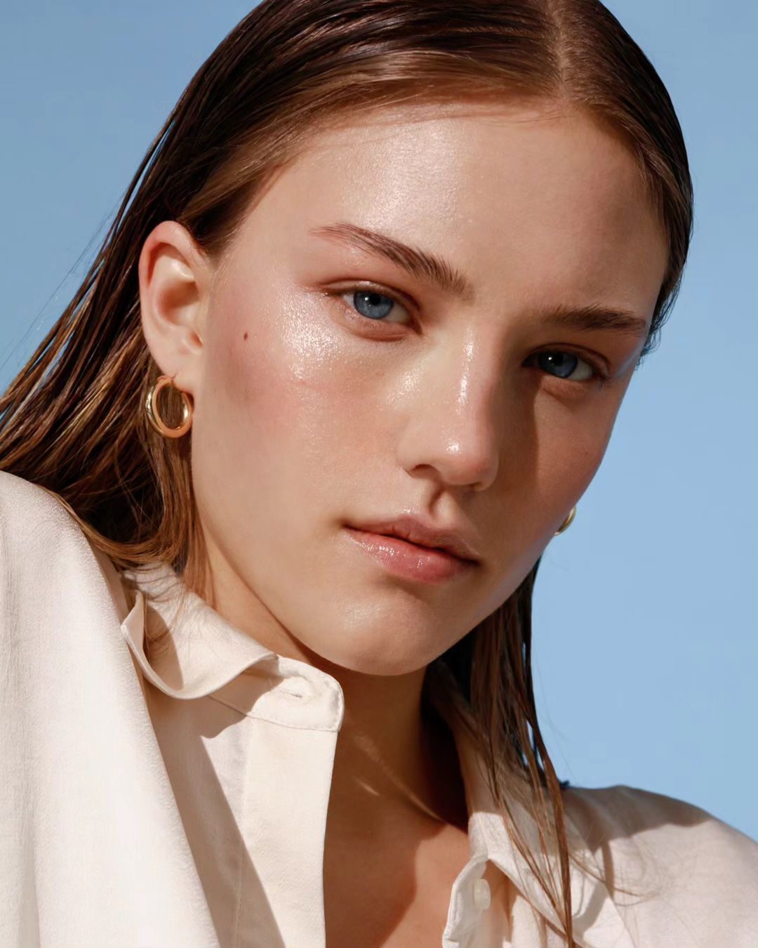 Close-up portrait of a fair-skinned woman with wet-look hair, gold hoop earrings, and a cream-colored top against a light blue background.
