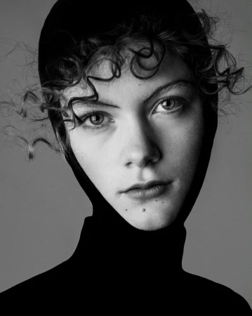 Close-up portrait of a woman wearing a black hood and turtleneck against a plain grey background by Christine Lutz