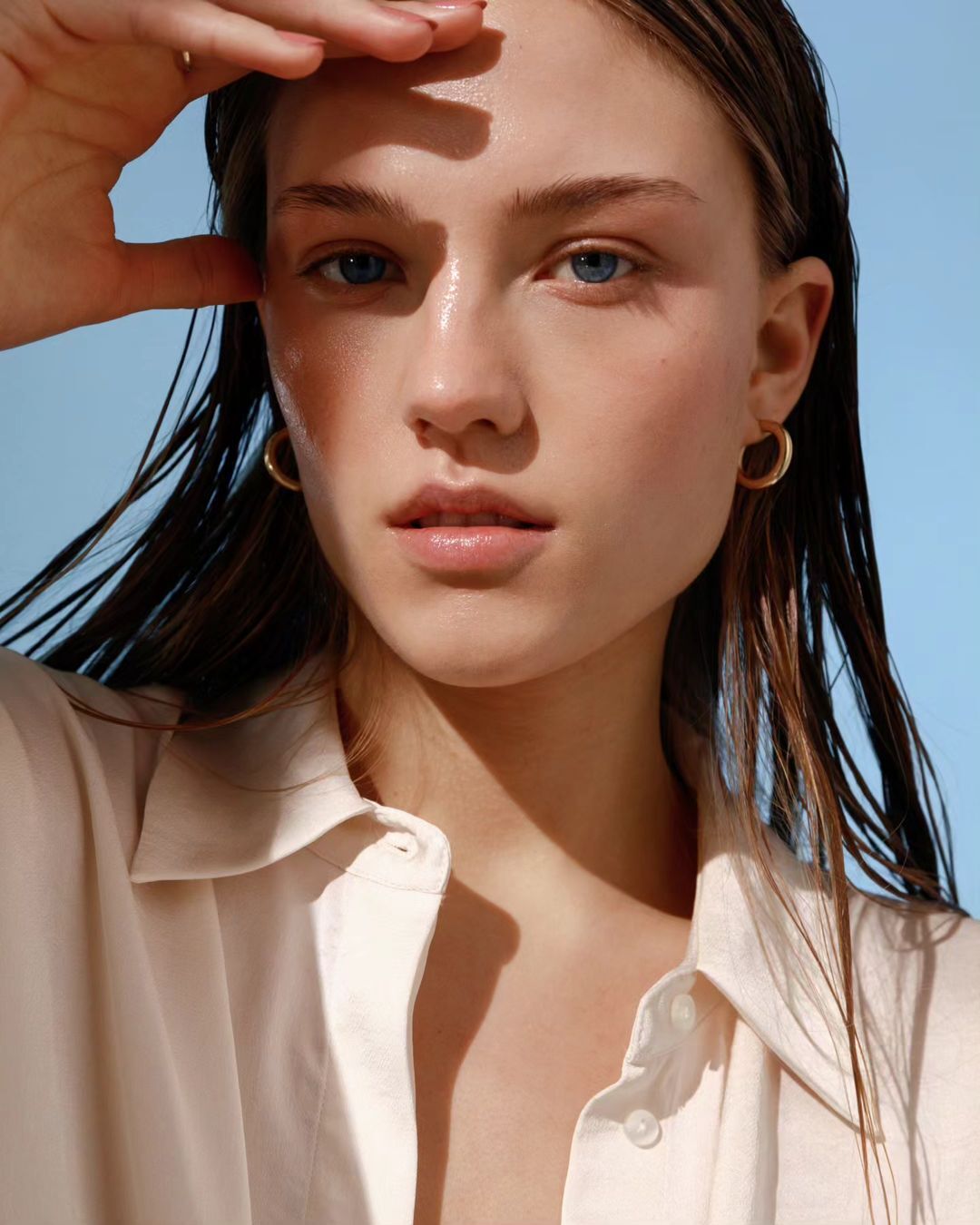 Close-up portrait of a young woman with wet brunette hair, wearing a cream-colored shirt and gold hoop earrings against a pale blue backdrop.
