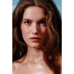 Close-up portrait of a woman with long brown wavy hair and a neutral expression against a clear blue sky background.