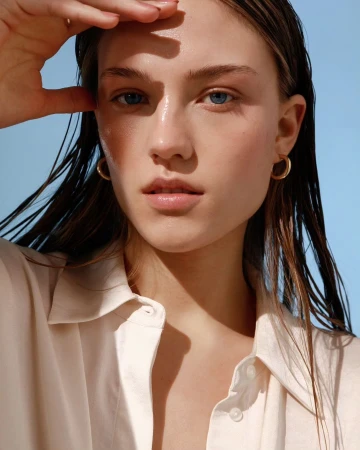 Close-up portrait of a woman with wet hair wearing a cream shirt and gold hoop earrings against a clear blue sky background.