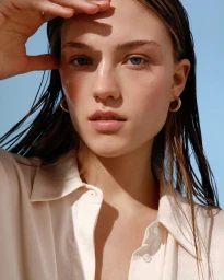 Close-up portrait of a woman with wet hair wearing a cream shirt and gold hoop earrings against a clear blue sky background.