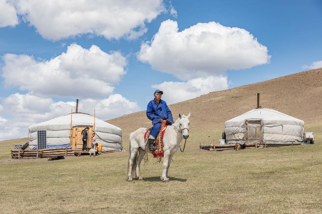 Mongolian man in traditional dress rides a white horse across grassland in front of two gers under a bright blue sky with fluffy clouds.