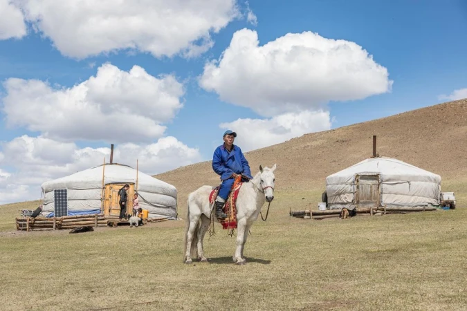 Mongolian man in traditional dress rides a white horse across grassland in front of two gers under a bright blue sky with fluffy clouds.