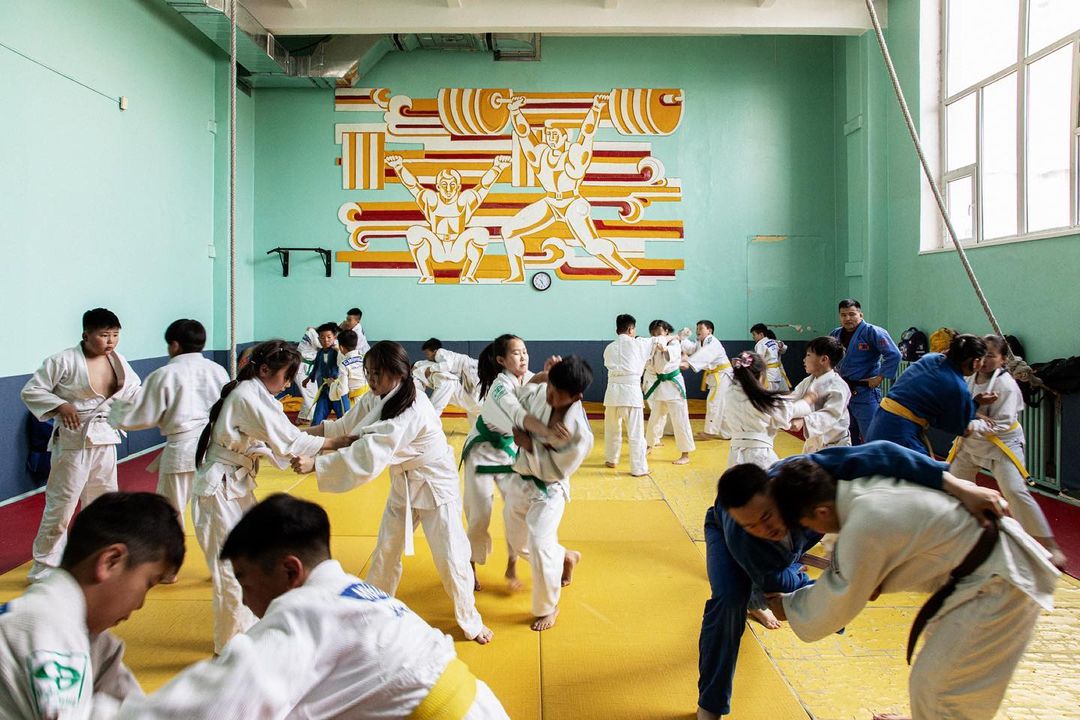 Indoor judo class with children in white judogis practicing throws, supervised by two instructors in blue judogis on a yellow floor.