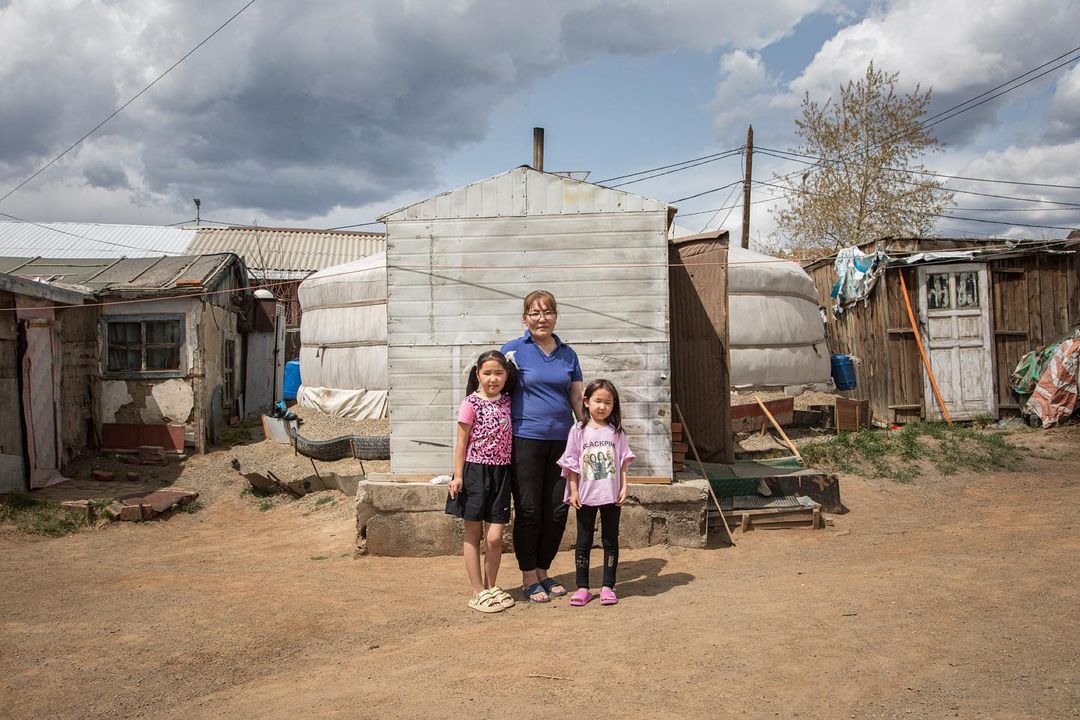 Family portrait: Woman with two young girls stand in front of a traditional yurt dwelling in a rural outdoor setting on a cloudy day.