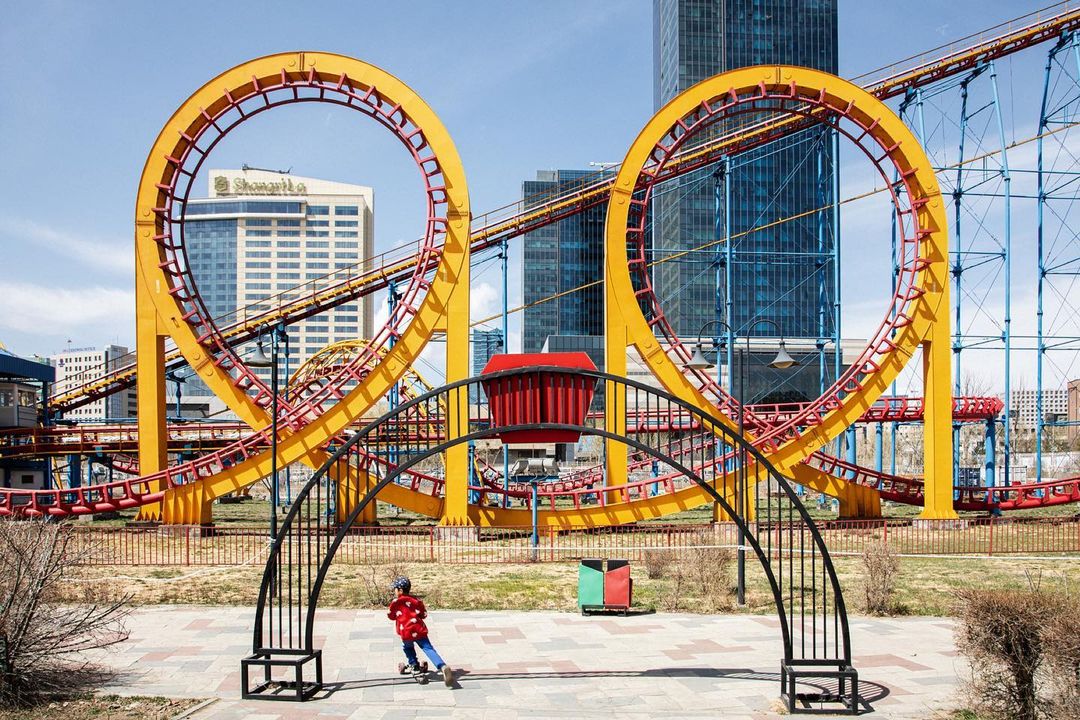 A child on a skateboard passes under a black metal arch in front of a yellow and red roller coaster with city buildings in the background.