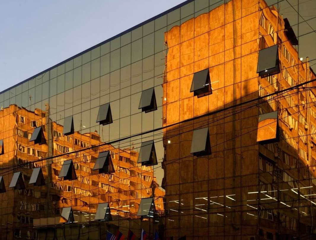 Building reflects off glass facade of modern building with open windows. Cityscape and light reflections create warm urban abstract.