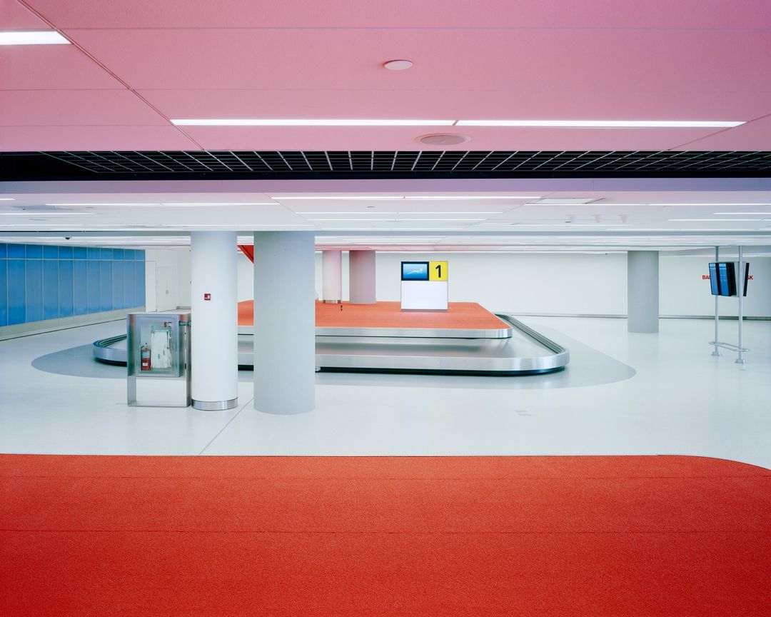 Empty airport terminal with baggage carousel, blue accent wall, and pale pink ceiling in a spacious, brightly lit interior.