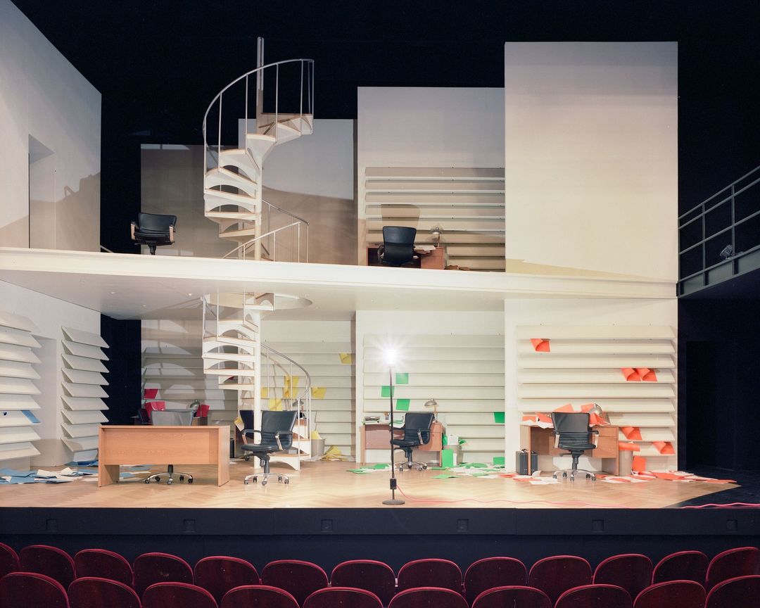 A multi-level stage set features office spaces, a spiral staircase, desks, chairs, and file cabinets under stage lighting, viewed from the audience.