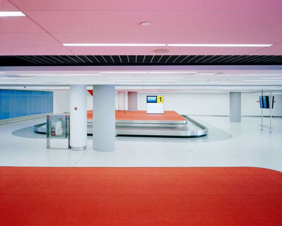 Modern airport interior with baggage claim carousel, pink-lit ceiling, blue accent wall, gray columns, and orange flooring.