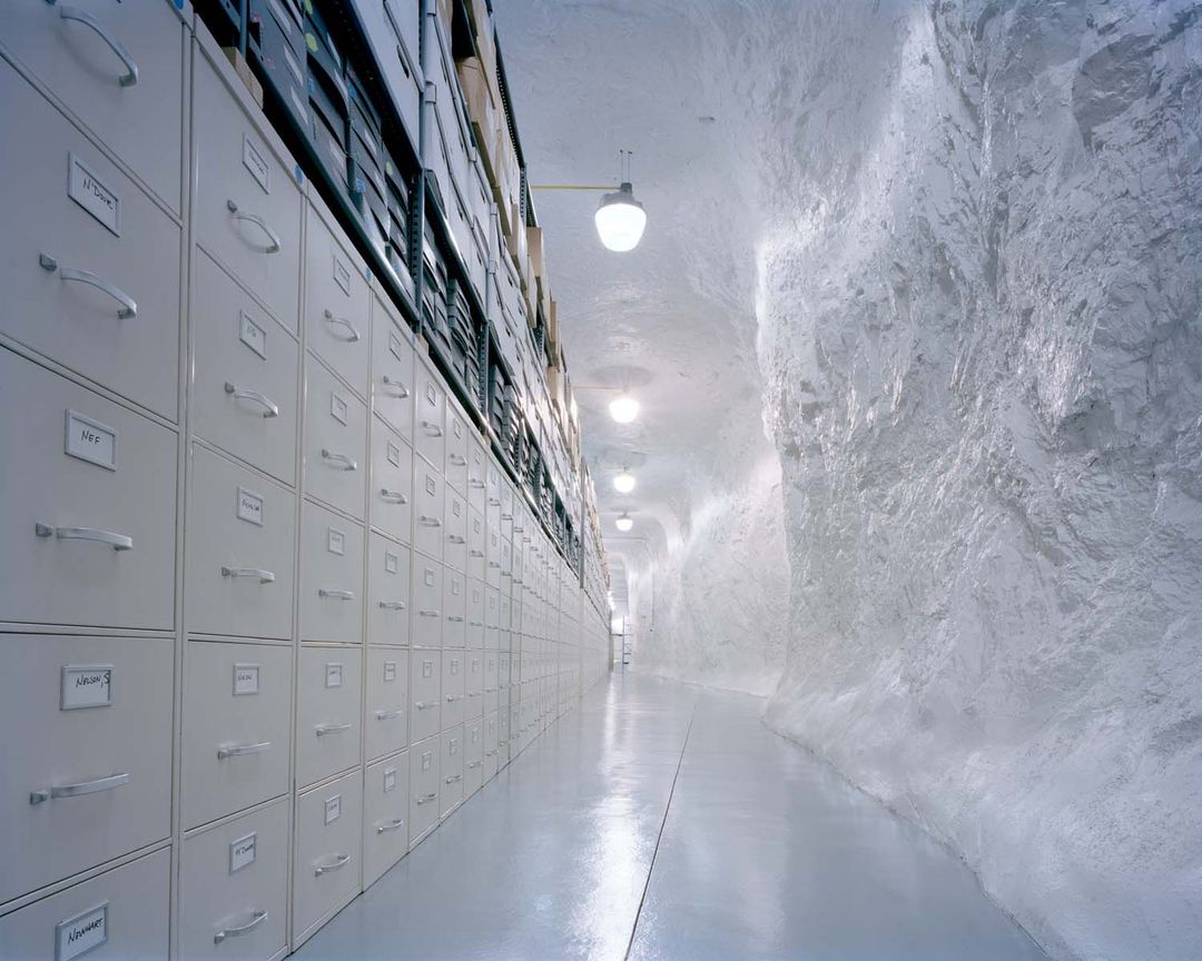 Long corridor with rows of metal filing cabinets receding into the distance against a textured white rock wall in an indoor setting.
