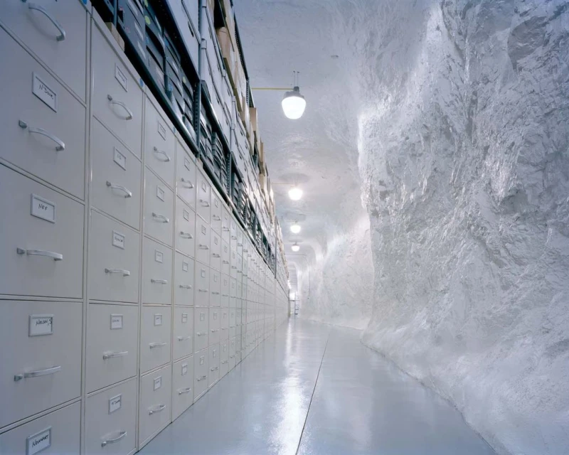 Long corridor with rows of metal filing cabinets receding into the distance against a textured white rock wall in an indoor setting.
