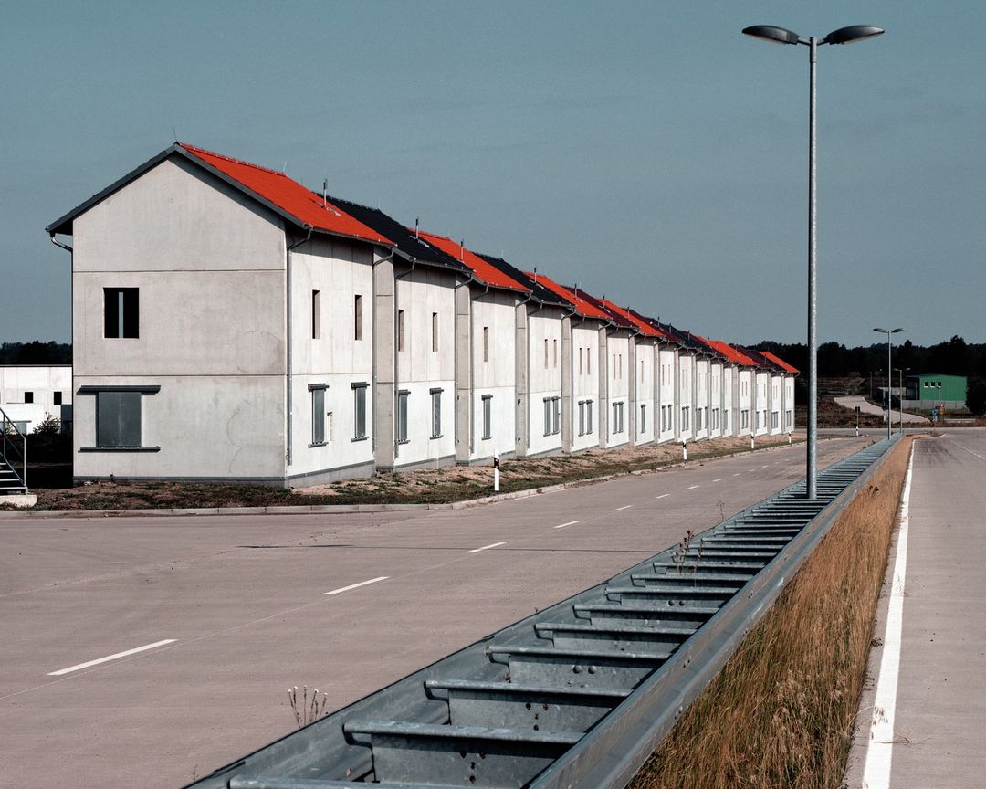 Row of identical two-story concrete houses with orange roofs stretches along a road under a blue sky, captured in a daylight exterior shot.
