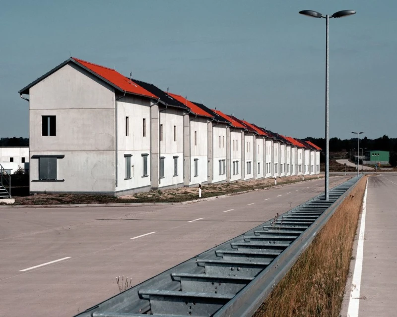 Row of identical two-story concrete houses with orange roofs stretches along a road under a blue sky, captured in a daylight exterior shot.