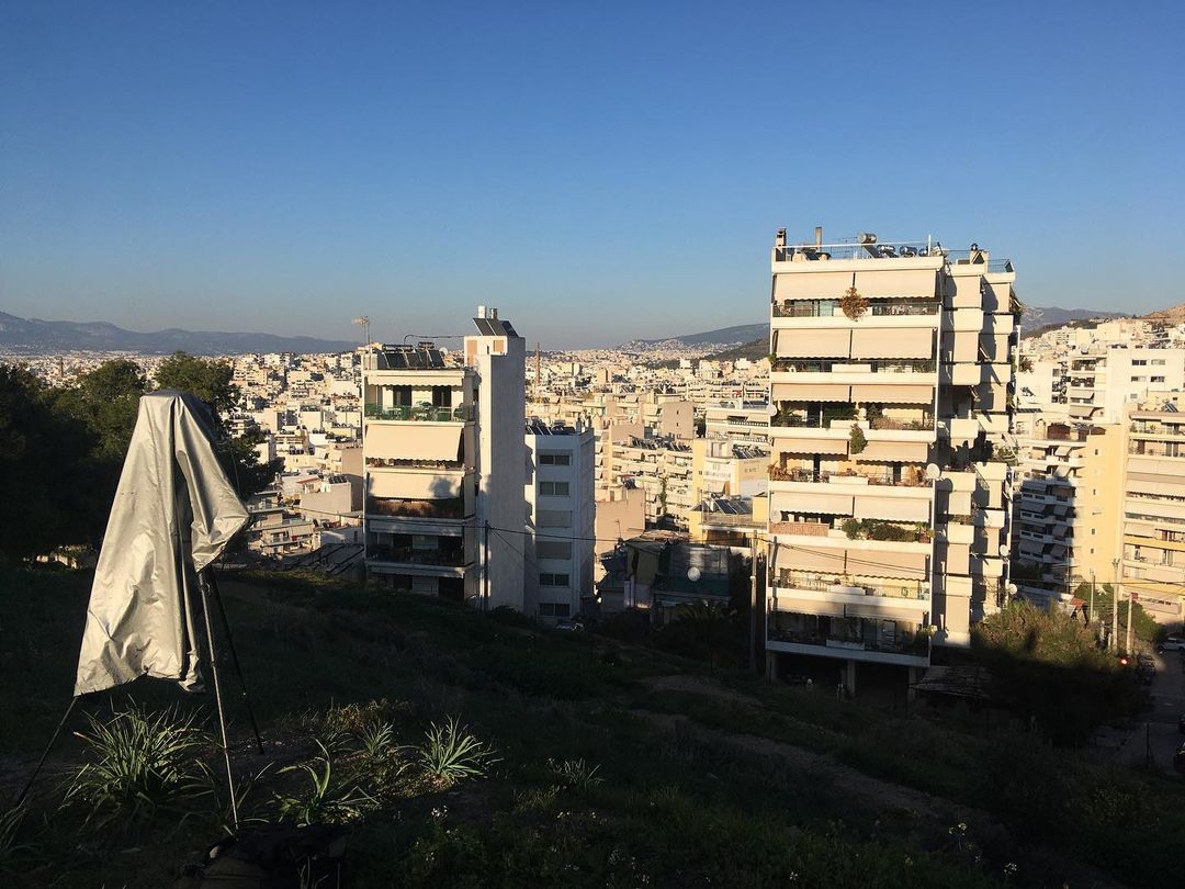Cityscape view with modern buildings, a hillside, and a reflector in the foreground under a clear blue sky on a sunny day.