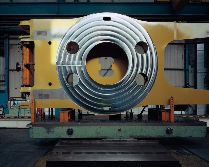 Close-up of a large yellow industrial machine with a circular silver metal component with holes in the center in an industrial environment.