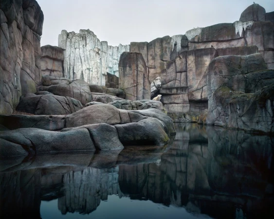 A polar bear sits in a constructed rock habitat with dark water in the foreground under an overcast sky, creating a contemplative scene.