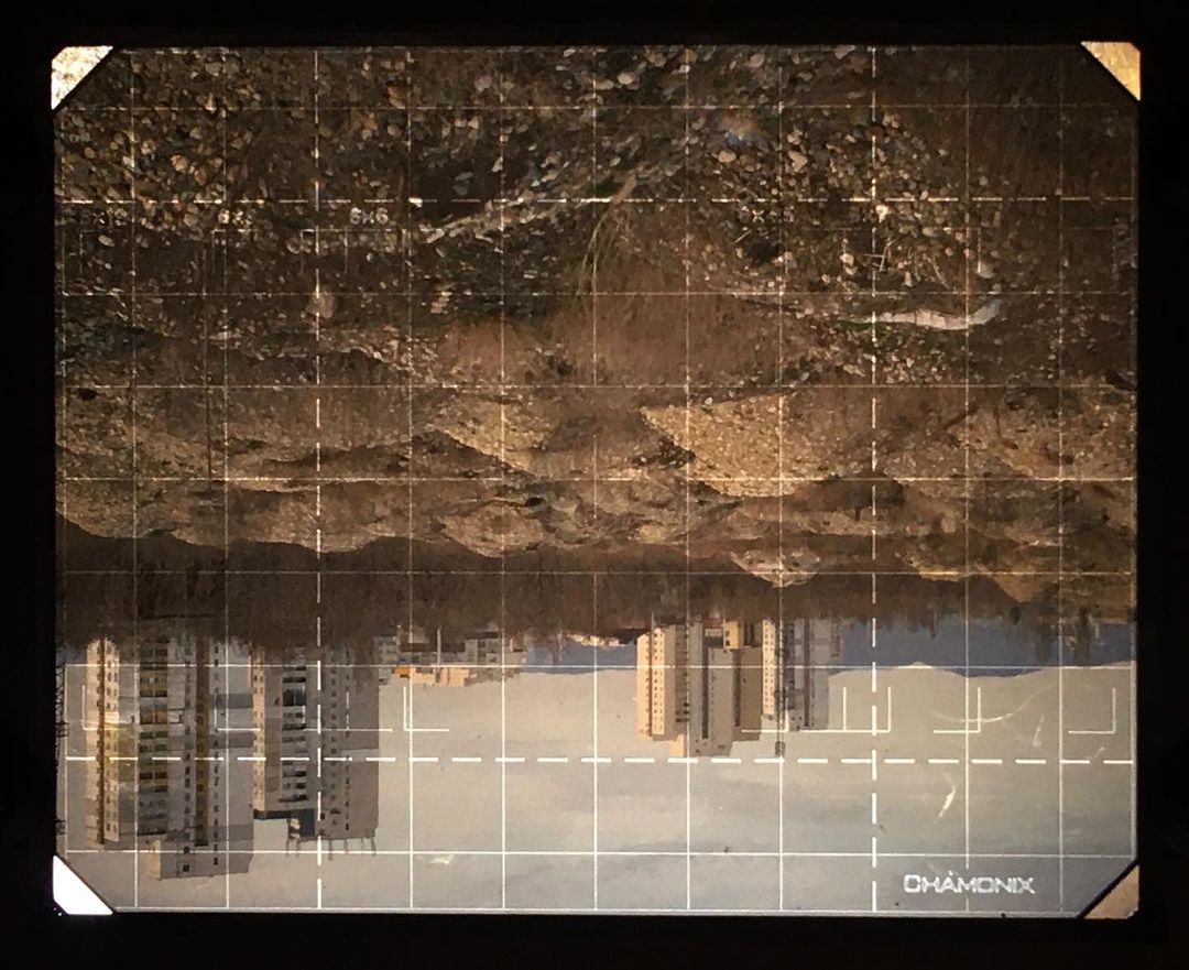 Inverted large-format camera viewfinder glass displaying an upside-down urban landscape with tall buildings and ground texture.
