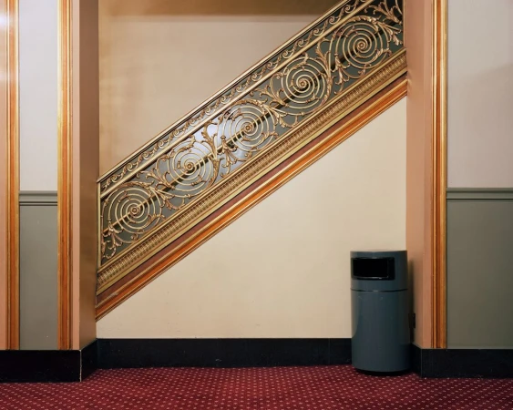 Interior view of decorative wrought iron staircase railing with gray trash receptacle on red patterned carpet in a hallway.