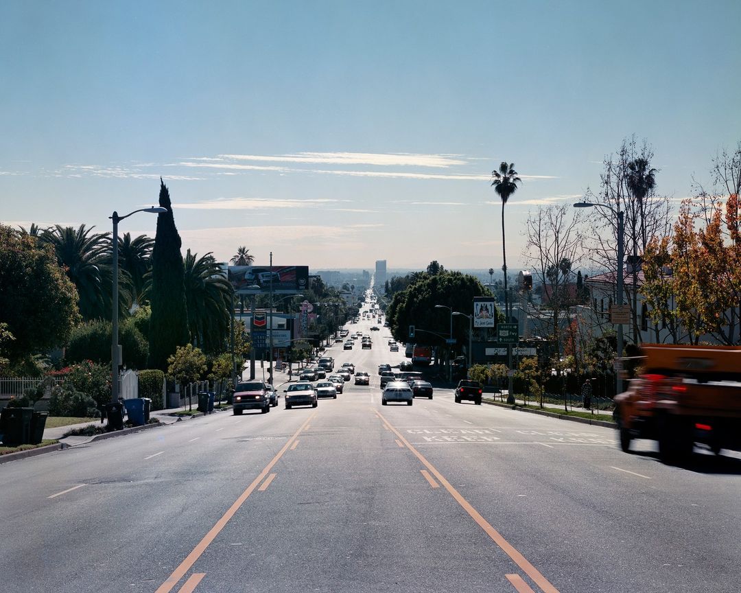 Cityscape view of a road lined with trees and cars on a sunny day, leading toward distant buildings and clear blue sky with light clouds.