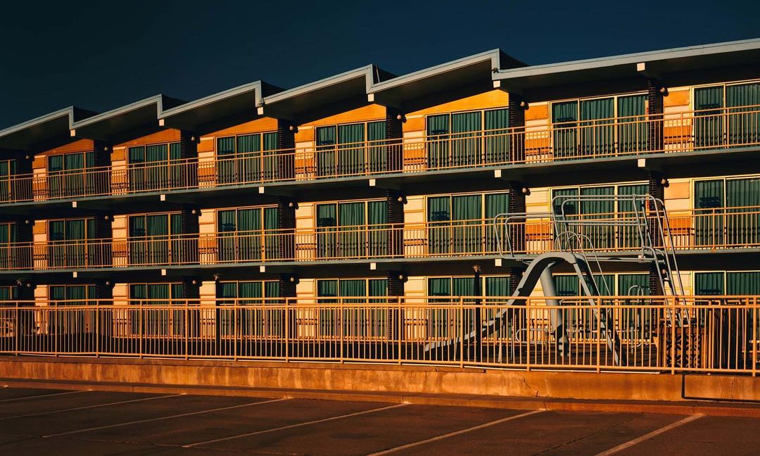 Exterior of a motel building with a yellow-painted facade, multiple stories, and balconies with metal railings, photographed in warm light.