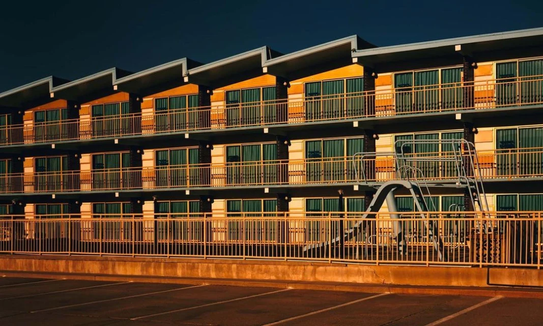 Exterior of a motel building with a yellow-painted facade, multiple stories, and balconies with metal railings, photographed in warm light.