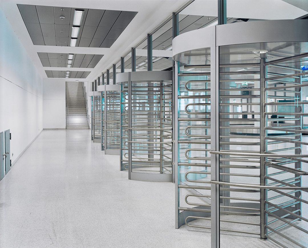 Interior view of a sterile modern commercial or institutional space with turnstiles in a row and a flight of stairs in the background.