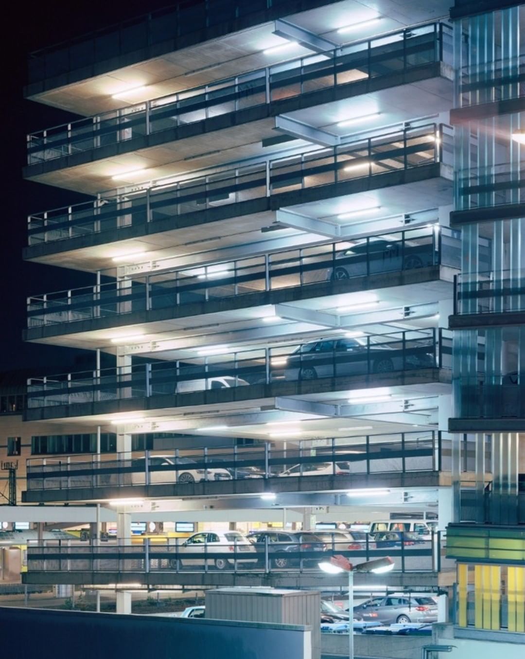 Exterior of a brightly lit parking garage at night with multiple parked cars on several levels, creating a structured, urban scene.
