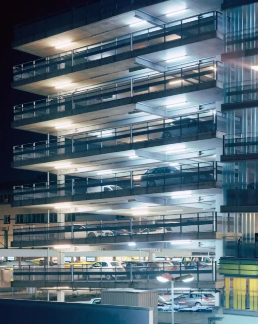 Exterior of a brightly lit parking garage at night with multiple parked cars on several levels, creating a structured, urban scene.