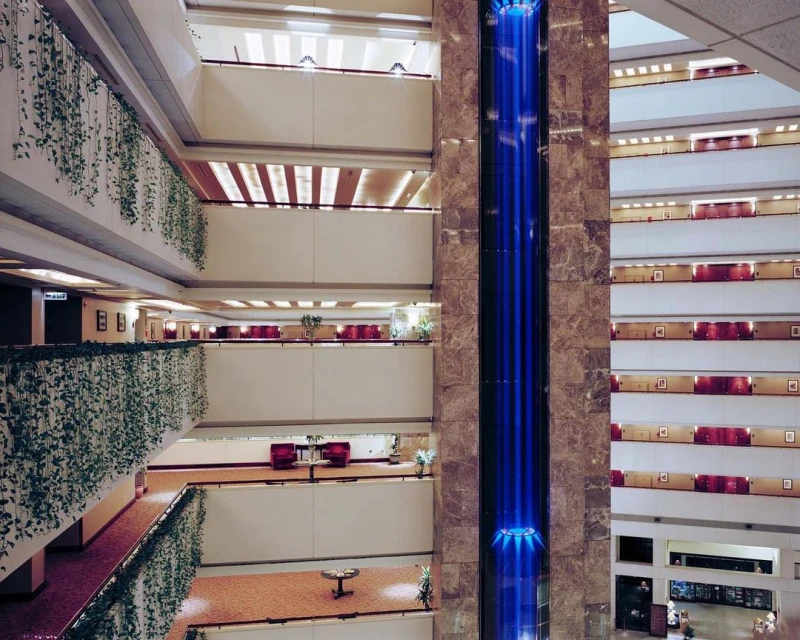 Indoor view of a multi-story hotel atrium featuring decorative greenery, a glass elevator, and multiple levels of rooms with balconies.