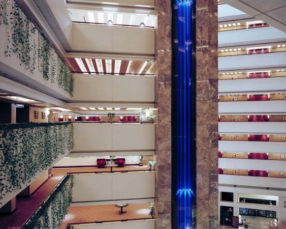 Indoor view of a multi-story hotel atrium featuring decorative greenery, a glass elevator, and multiple levels of rooms with balconies.