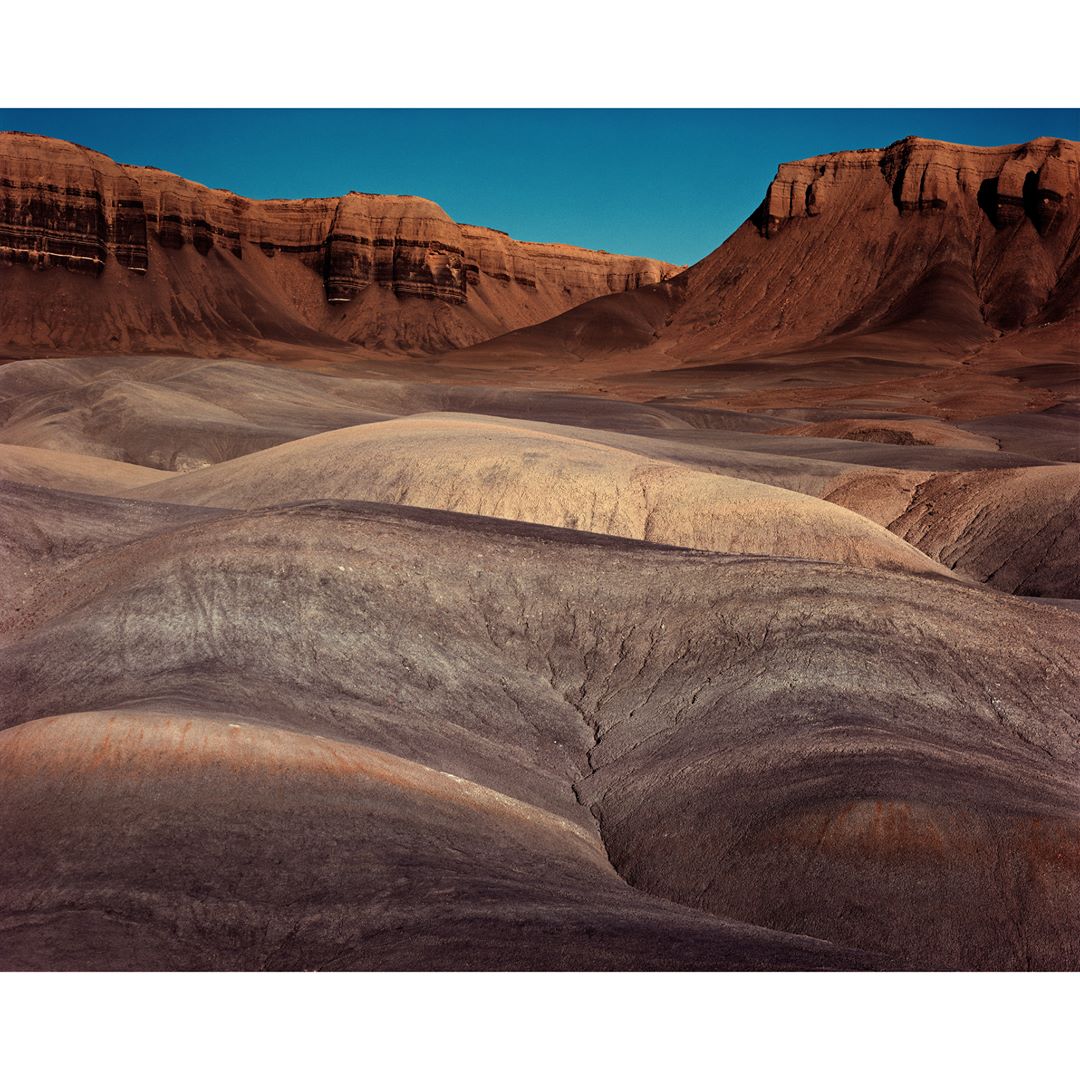 Vast desert landscape featuring eroded rolling hills in shades of brown and grey, set beneath a clear blue sky with mountain range in the background.