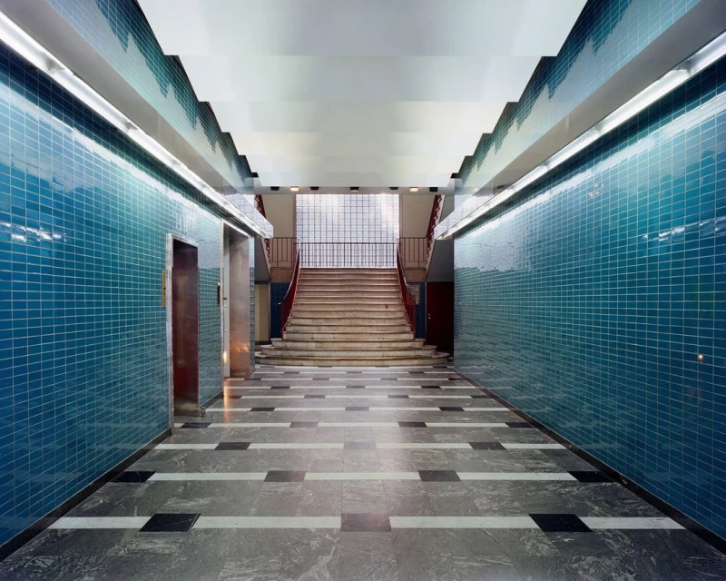 Interior view of a hallway with blue tile walls, a gray tile floor with black and white accents, stairs, and elevator doors.