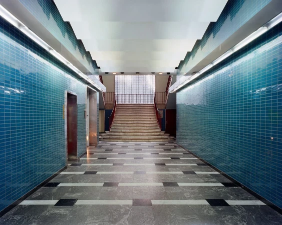 Interior view of a hallway with blue tile walls, a gray tile floor with black and white accents, stairs, and elevator doors.