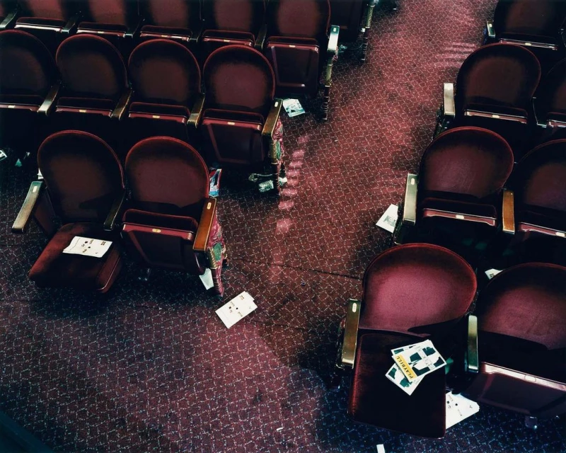 Overhead shot showcases rows of unoccupied red velvet theater seats with scattered papers and debris on the patterned maroon carpeted floor.