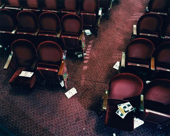 Overhead shot showcases rows of unoccupied red velvet theater seats with scattered papers and debris on the patterned maroon carpeted floor.