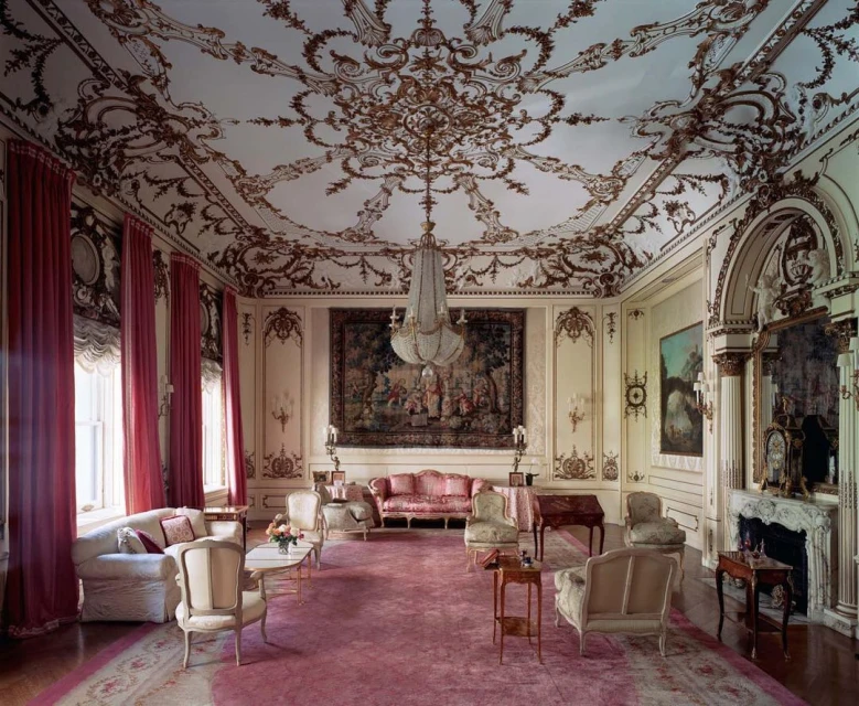 Ornate living room with high decorative ceiling, antique furniture, pink carpet, chandelier, and pink drapes in a traditional style.