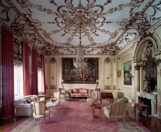 Ornate living room with high decorative ceiling, antique furniture, pink carpet, chandelier, and pink drapes in a traditional style.