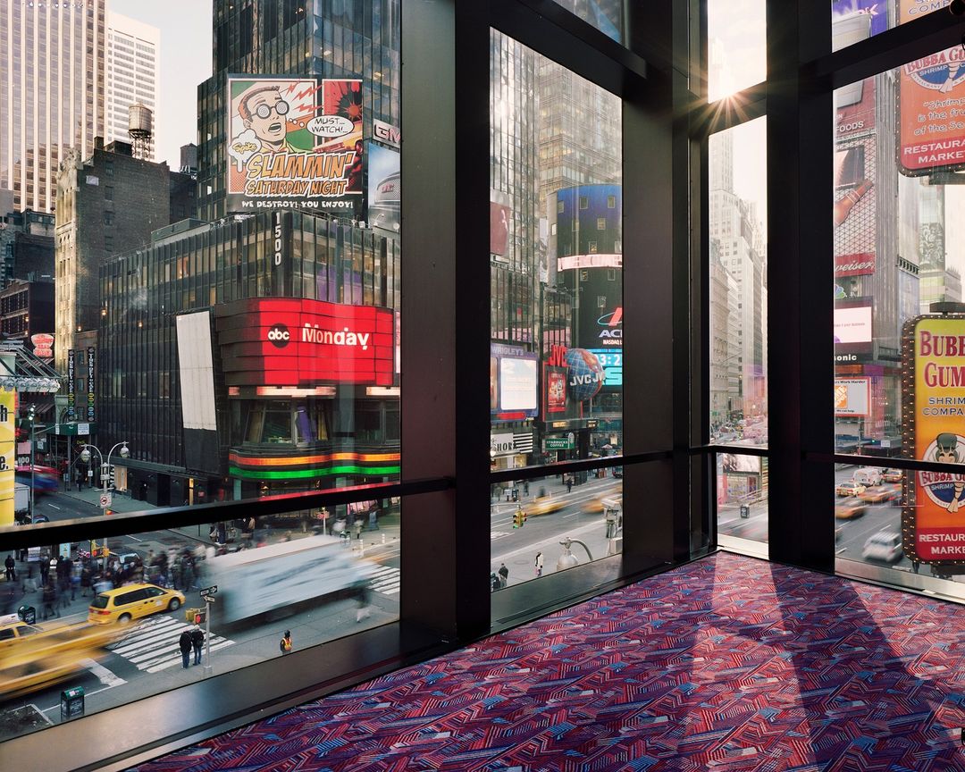 Cityscape view from inside building overlooks Times Square, featuring city streets, billboards, and architecture on a sunny day.