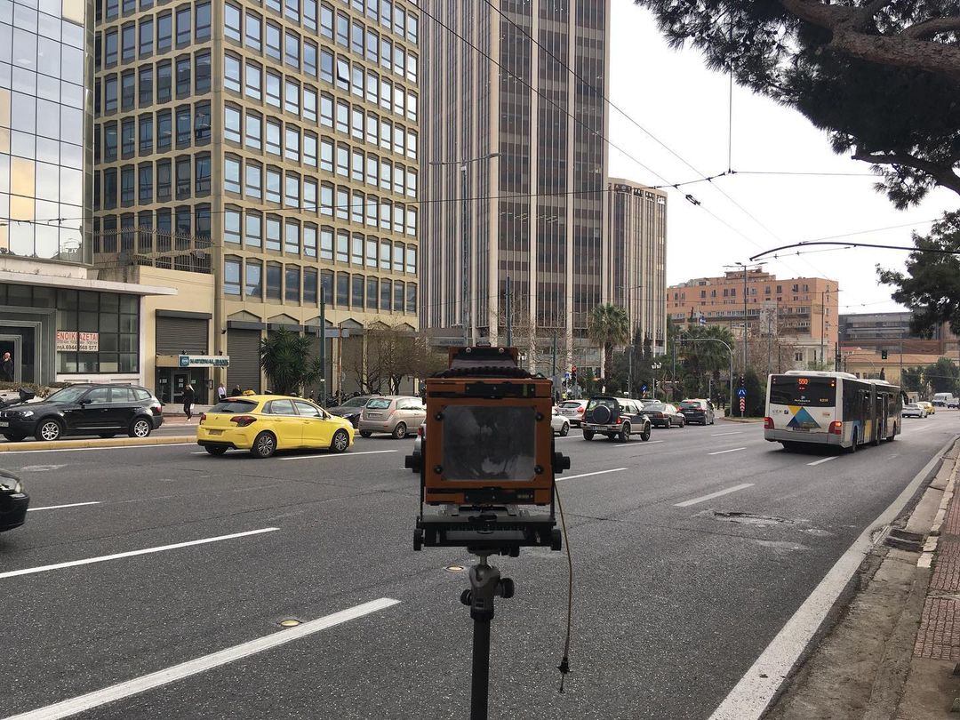 Large format camera on tripod in the middle of a city street with traffic and tall buildings in the background on a cloudy day.