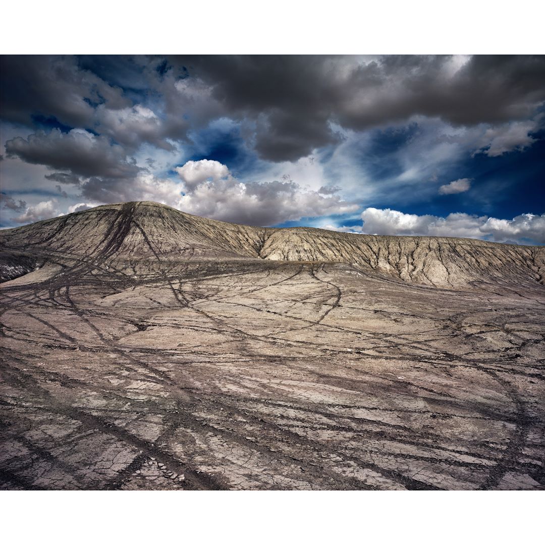 Arid landscape featuring a large hill with eroded gullies under a dramatic cloudy sky with dark tire tracks crisscrossing the foreground.